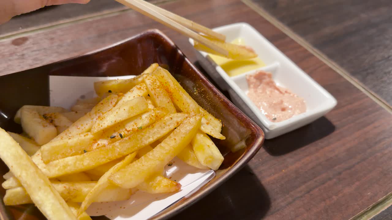 Hand uses chopsticks to dip seasoned French fry in mayonnaise sauce under warm restaurant lighting