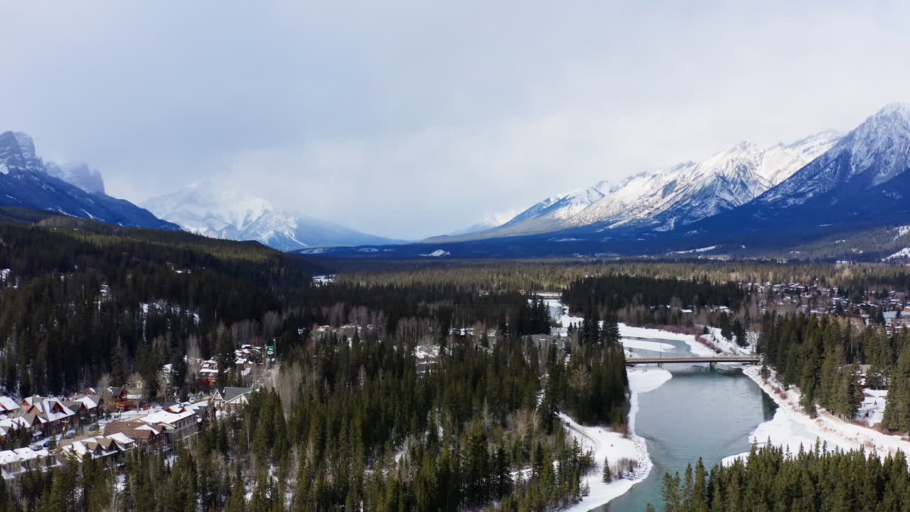 A drone glides over Canmore, capturing a snow-covered river, dense forest, and dramatic mountain peaks in the BANFF range.