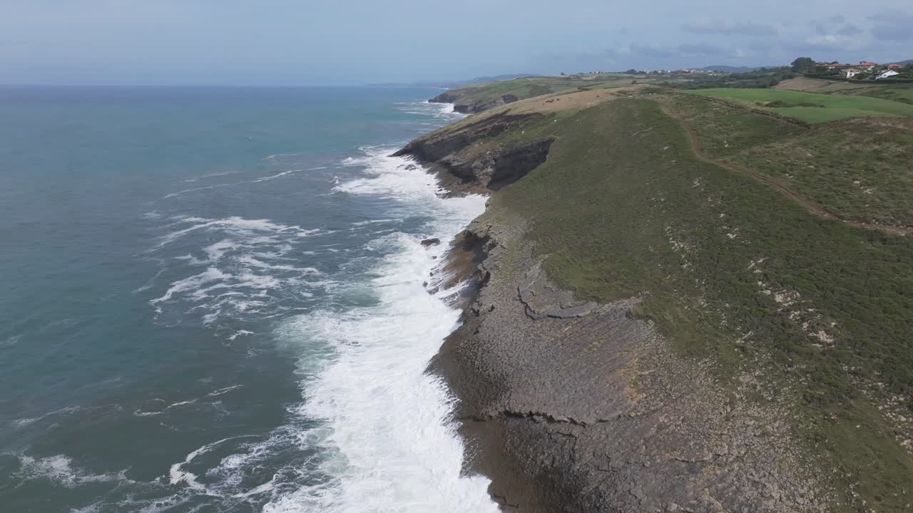 A sideways flight leaves the green meadows and cliffs of the Cantabrian coast to fly over the open sea. A dividing line shows land on one side and water on the other; waves crash against the rocks