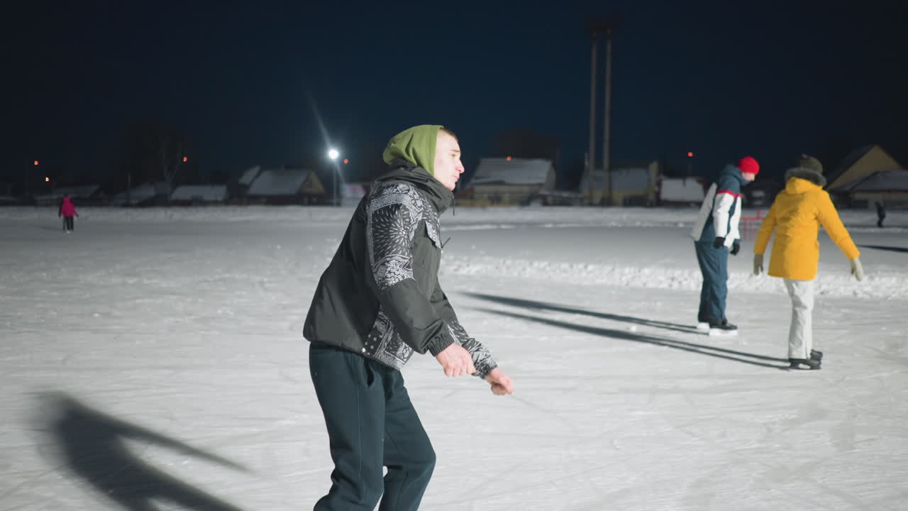 young people skating on well-lit outdoor ice rink during night in winter as others glide in background wearing winter clothing under artificial lighting with visible shadows on ice surface