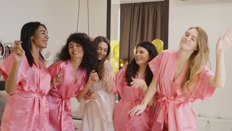 grupo de amigas multiétnicas y novias con camisones de seda rosa y blanca mientras bailan en una habitación en una reunión nupcial