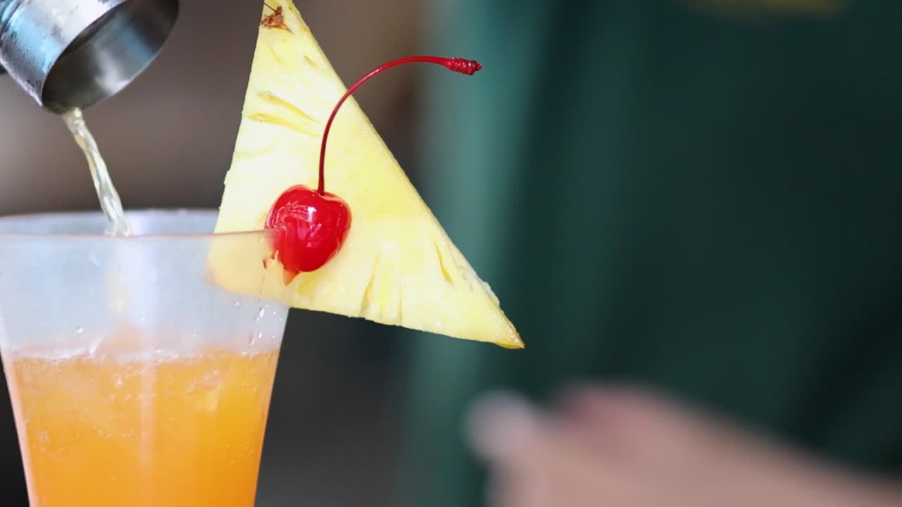 A close-up of a bartender pouring an orange cocktail, garnished with pineapple and cherry.