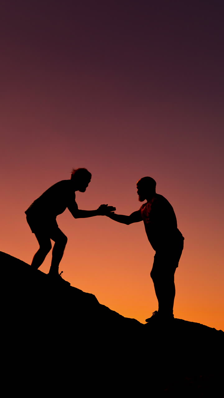 Silhouettes of Two People Helping Each Other Climb a Hill at Sunset