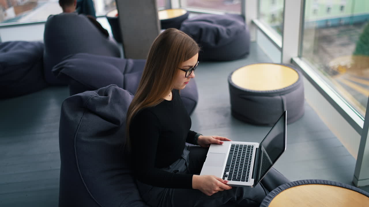Attractive woman wearing glasses works on a laptop. Young businesswoman working on a laptop in office. Female professional with gadget near the window at office. Top view.