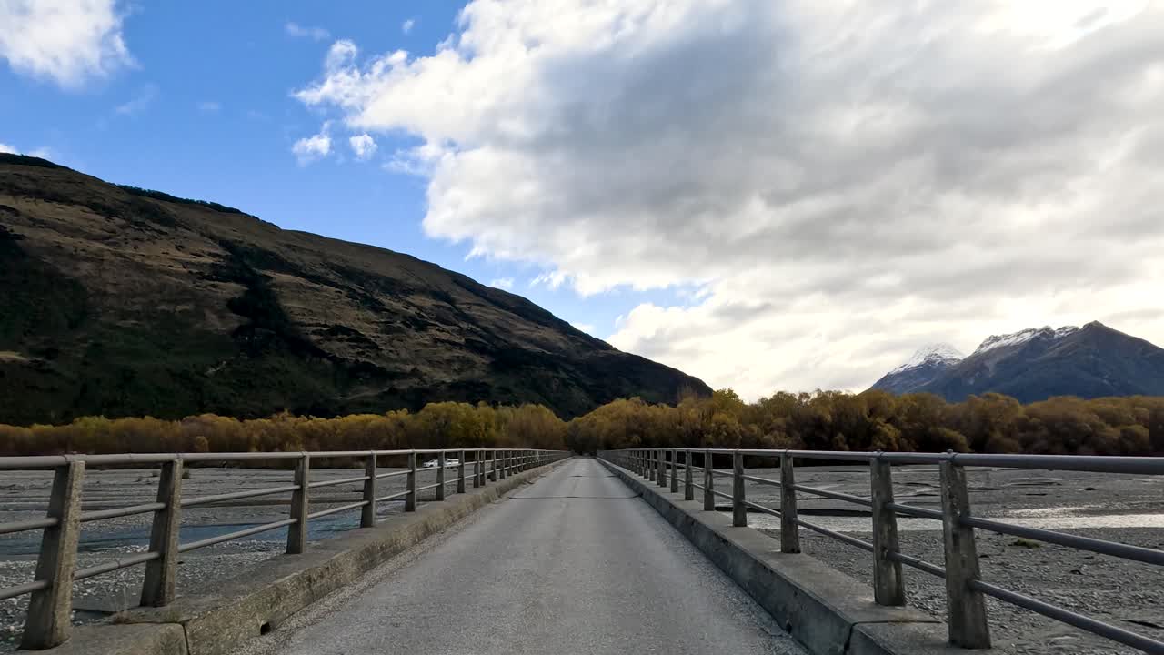 Vehicle drives over rural bridge with river, autumn trees, and mountains under partly cloudy sky