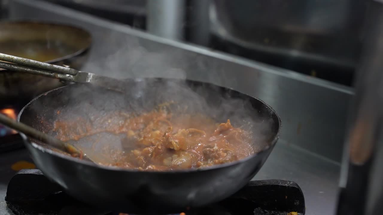 Uno chef mescola pollo karahi fumante in un wok nero su fiamma viva in una cucina di ristorante.