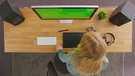 overhead view of female graphic designer or photo retoucher putting on wireless headphones before sitting at desk with graphics tablet working at computer with green screen in creative office - shot in slow motion