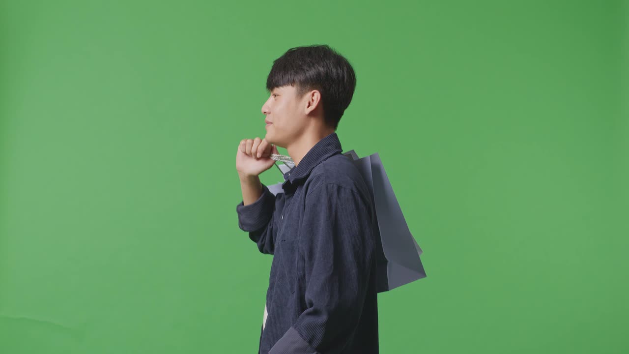 Side View Of An Asian Shopping Man With Shopping Bags Smile And Walking In Front Of Green Screen