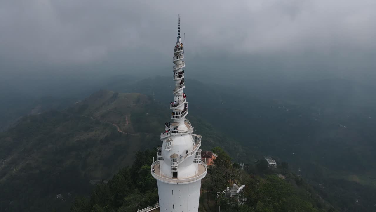 Several tourists stand close together on the white narrow spiral stairs on the Ambuluwawa tower in Sri Lanka to enjoy the beautiful view of the green nature on a cloudy day. Wide drone panning shot