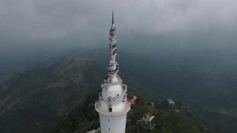 several tourists stand close together on the white narrow spiral stairs on the ambuluwawa tower in sri lanka to enjoy the beautiful view of the green nature on a cloudy day. wide drone panning shot