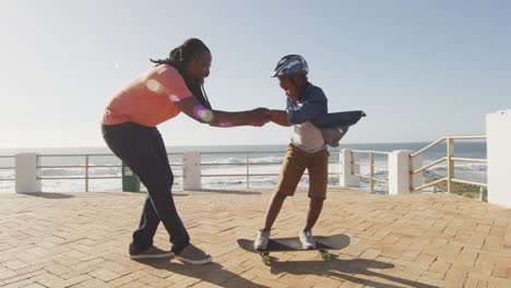 video de un feliz padre afroamericano aprendiendo a su hijo a patinar en el paseo marítimo