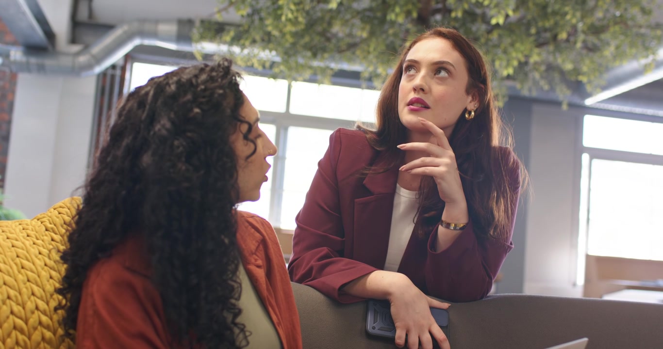 Collaboratori diversi: una donna seduta con un top color ruggine digita al laptop mentre un collega si china, avviando un aggiornamento del telefono.