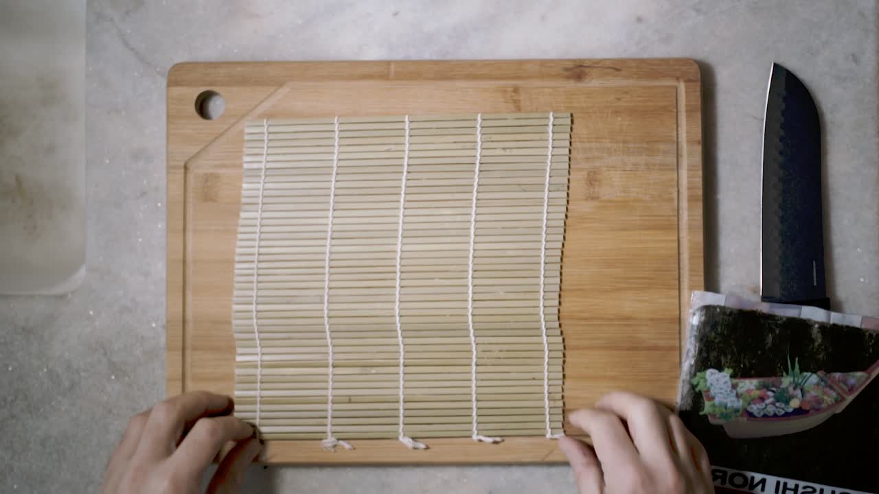 Top down view on kitchen counter with a mat for Sushi rice with two hands adjusting and lying it in position in preparation to make a Sushi roll