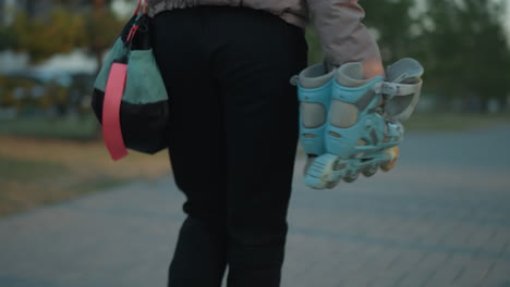 a girl, dressed in a peach jacket and black trousers, carries a pair of rollerblades and a gym bag as she walks through a park during sunset. focusing on the rollerblades and her casual attire