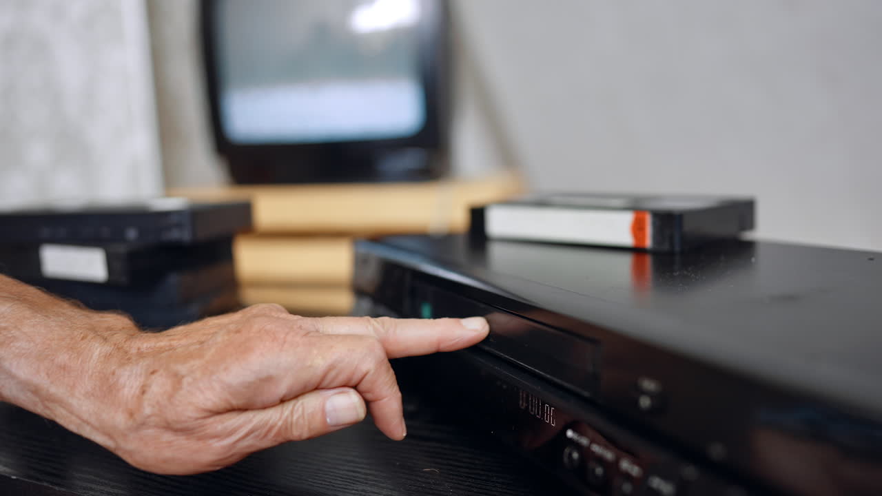 Hand inserting VHS tape into VCR. A person slides a VHS cassette into an old video recorder beside a blurred TV screen