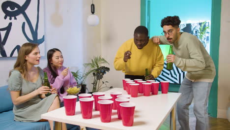 happy multiethnic young friends playing beer pong at home