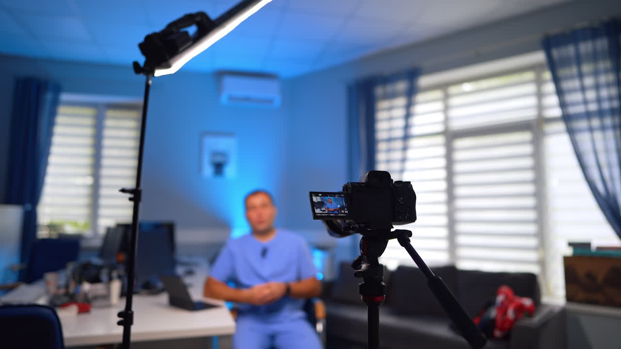Creazione di contenuti vlog medici. Un dottore in uniforme blu parla alla telecamera nel suo ufficio. Messa a fuoco selettiva