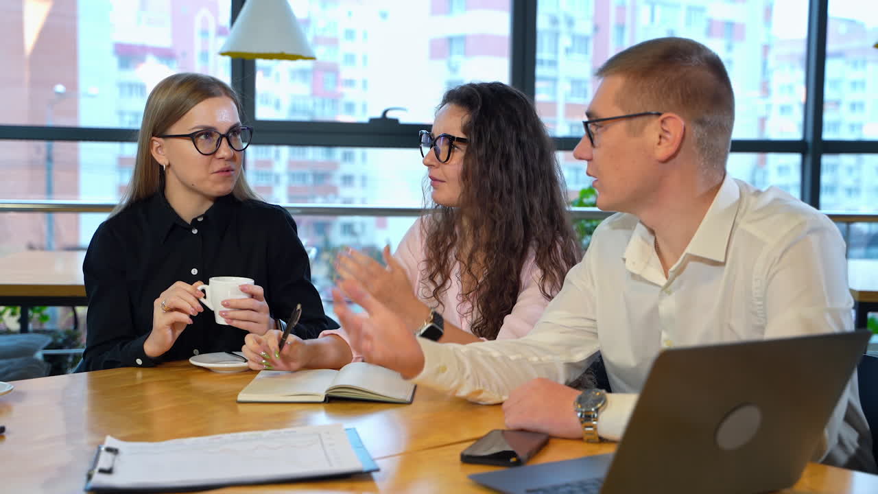 Young coworkers sit at the desk discussing something actively. People in glasses sharing ideas at work. Blurred cityscape at background.