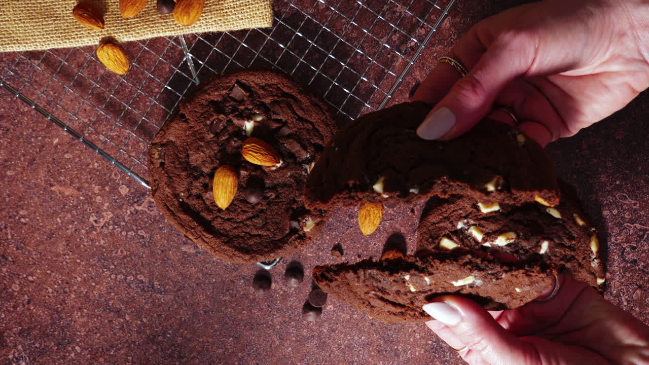 Vista dall'alto di mani che spezzano un morbido biscotto al cioccolato con mandorle e scaglie di cioccolato. La mollica e la consistenza appena sfornate sono rivelate su un tavolo rustico con una griglia di raffreddamento e ingredienti.