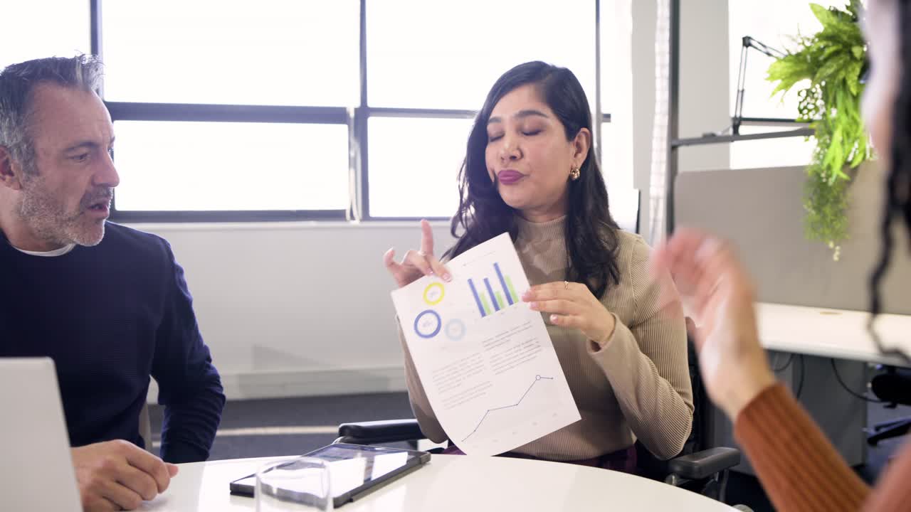 Mulher hispânica de meia-idade em mesa de reunião, levantando tablet que mostra gráficos e, em seguida, apresentando relatório