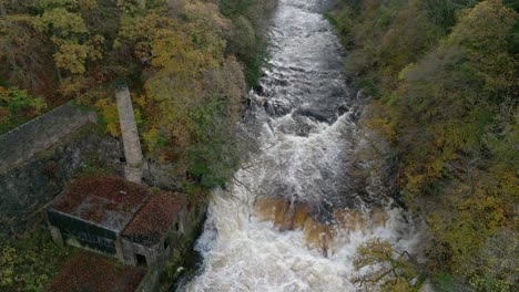 Drohnenaufnahmen-Direkt-über-Einem-Schnell-Fließenden-Fluss-Und-Wasserfall,-Umgeben-Von-Alten-Gebäuden,-Neigen-Sich-Nach-Oben,-Um-Dem-Fluss-Zu-Folgen,-Während-Herbstliche-Bäume-Die-Schlucht-überragen