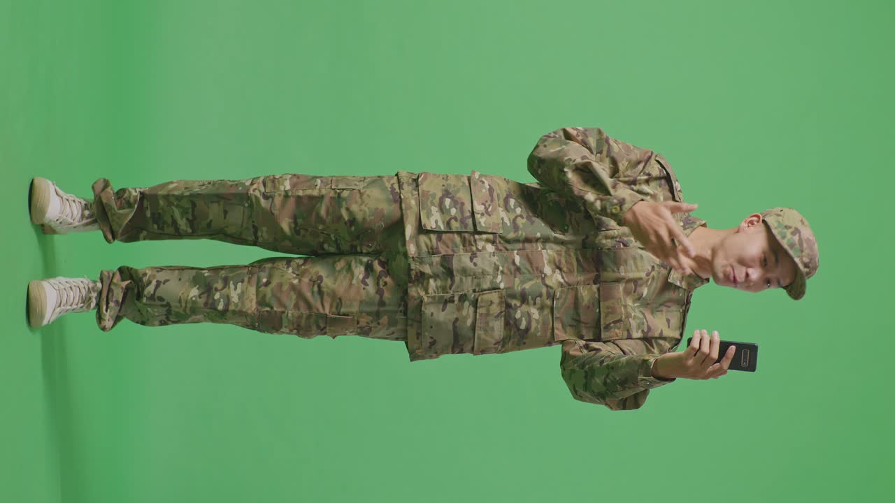 Full Body Of Asian Man Soldier Waving Hand Having A Video Call On Smartphone While Standing In The Green Screen Background Studio