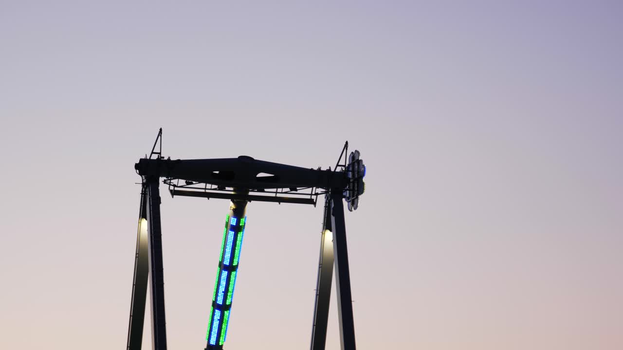 Wide shot of thrill seekers swinging back and forth on a giant Frisbee pendulum ride under a pastel sunset sky