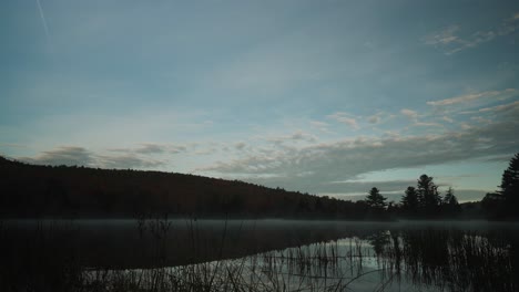 Fog-rolling-across-calm-glass-pond-as-sunrise-light-spreads-on-mountains-behind