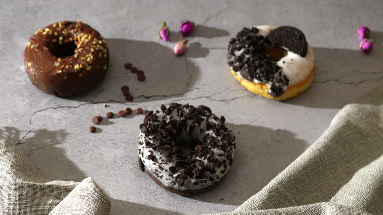 A hand with pink nails reaches out to grab a white-glazed cookie crumble donut from a rustic table. Surrounding it are chocolate chips, rosebuds, and other donuts in soft, natural light.