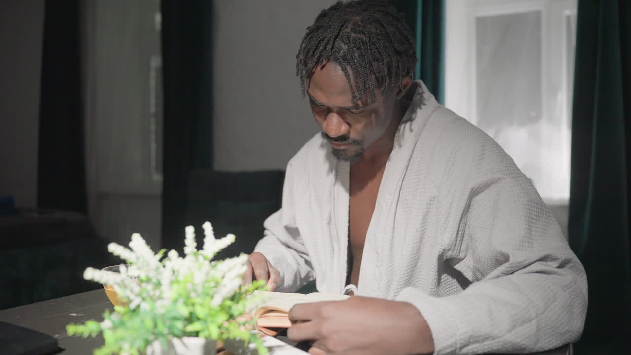 Champ enjoys tea while reading a book in peaceful home setting. Calm morning routine with cozy robe, bright natural light, and plant decor creating relaxing, mindful atmosphere for quiet moments