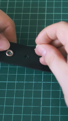 cu: skilled girl worker hands fix silver eyelets in leather belt on green cushion board in workshop close view