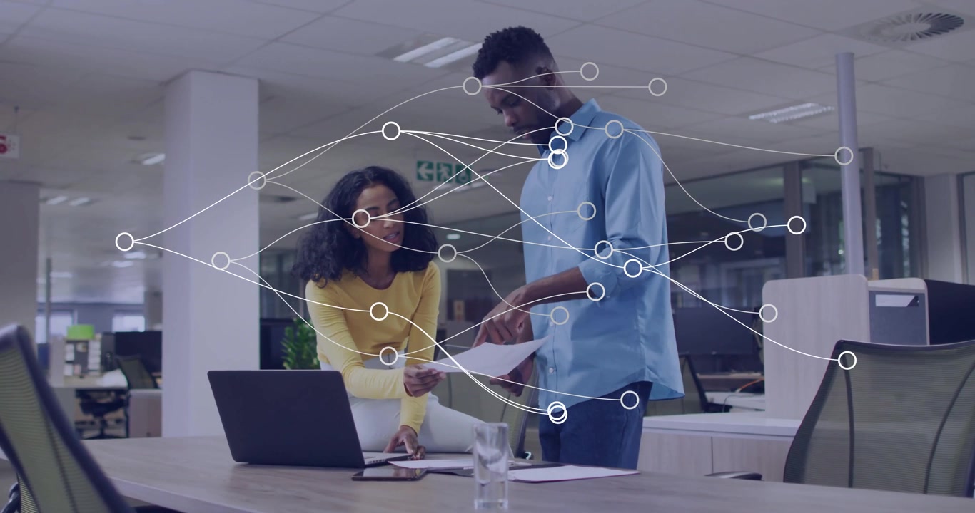 Woman tapping keys triggers digital nodes overlay, colleague examining document in business meeting