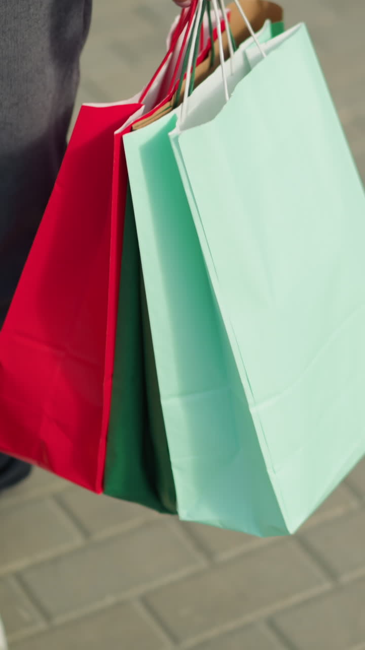 vista de la pierna de un individuo que usa corredores grises y zapatillas de deporte de lona blanca, sosteniendo bolsas de compras verdes, menta y rojas, caminando casualmente por un camino entrelazado en un entorno urbano