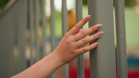 close-up of woman's hand with neatly polished nails gently gliding along iron fence bars, capturing the soft touch and elegance of her manicure in a reflective moment, with blurred background