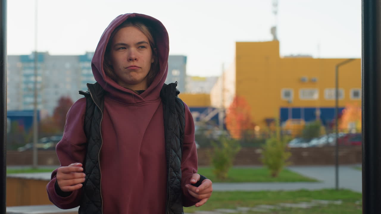Break moment when female athlete makes dance moves under pull up bar in urban park outdoor gym with burgundy hoodie vest rubber floor autumn foliage bench and city buildings in background