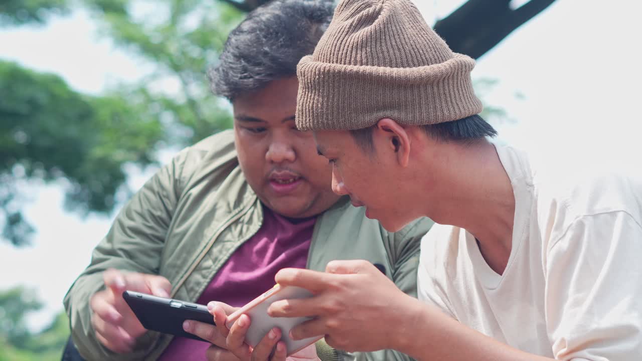 Two Young Asian Men Sits On Park Play Mobile Game On Smartphone Together, Clenched Fist, Celebrating Victory
