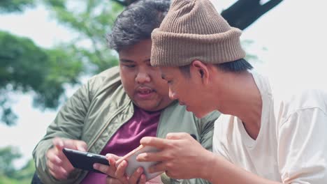 two young asian men sits on park play mobile game on smartphone together, clenched fist, celebrating victory