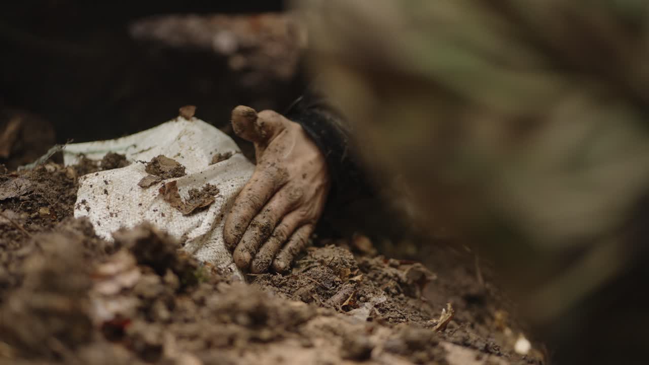 Close up of hooded, dirt covered man lying partially buried and unresponsive as a military character touches him, forest setting