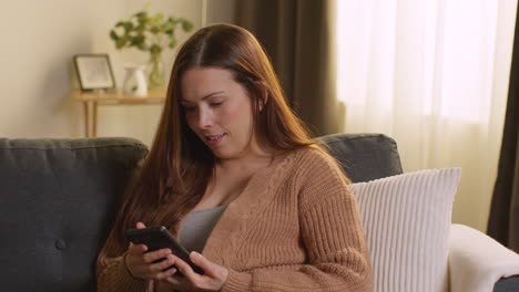 Laughing-Woman-Sitting-On-Sofa-At-Home-Using-Mobile-Phone-To-Check-Social-Media-Message-And-Scrolling-Online