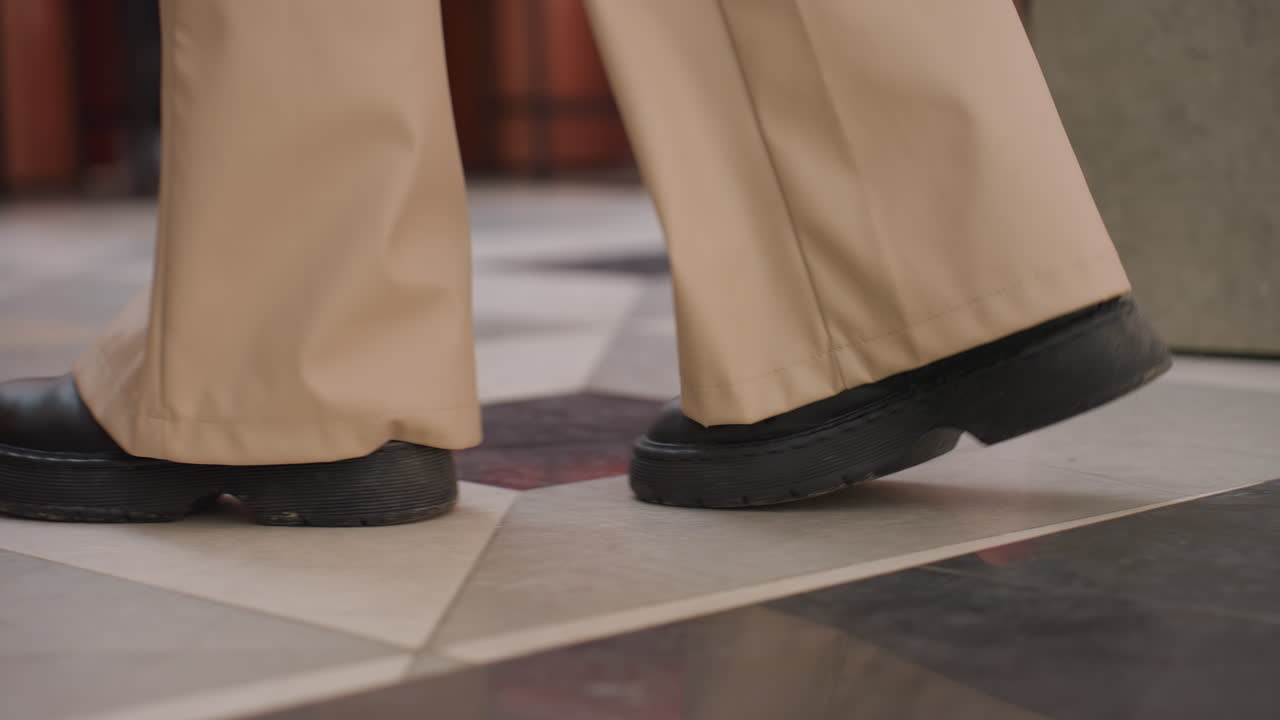 Leg view of person wearing black boots walking on well-tied floor toward decorative plant, urban structure in background, showcasing modern indoor environment with clean floor and stylish design