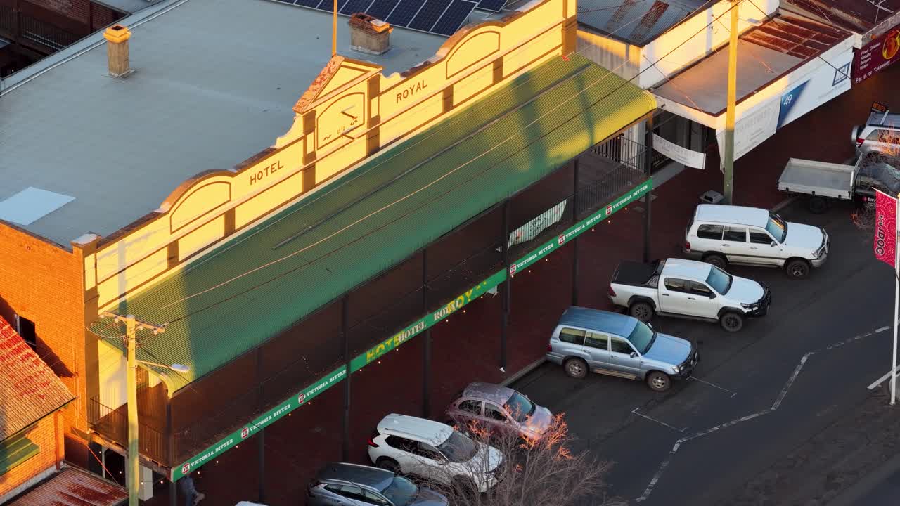Aerial view of a red semi truck driving past parked cars and a historic hotel at sunset in Coonabarabran, New South Wales. Warm, golden hour lighting with a slight downward camera angle