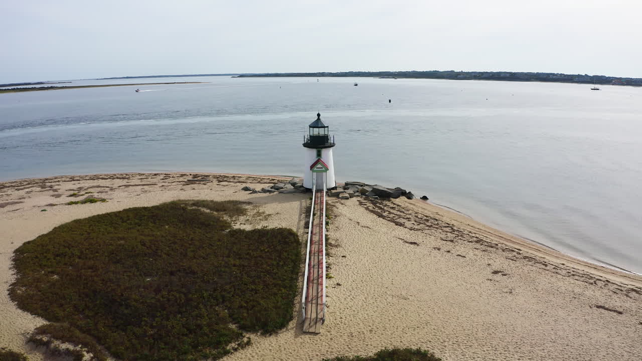 Vuelo cinematográfico de dron saliendo del Faro de Brant Point en Nantucket, Massachusetts, revelando la costa arenosa y las extensas aguas del puerto vistas desde arriba.
