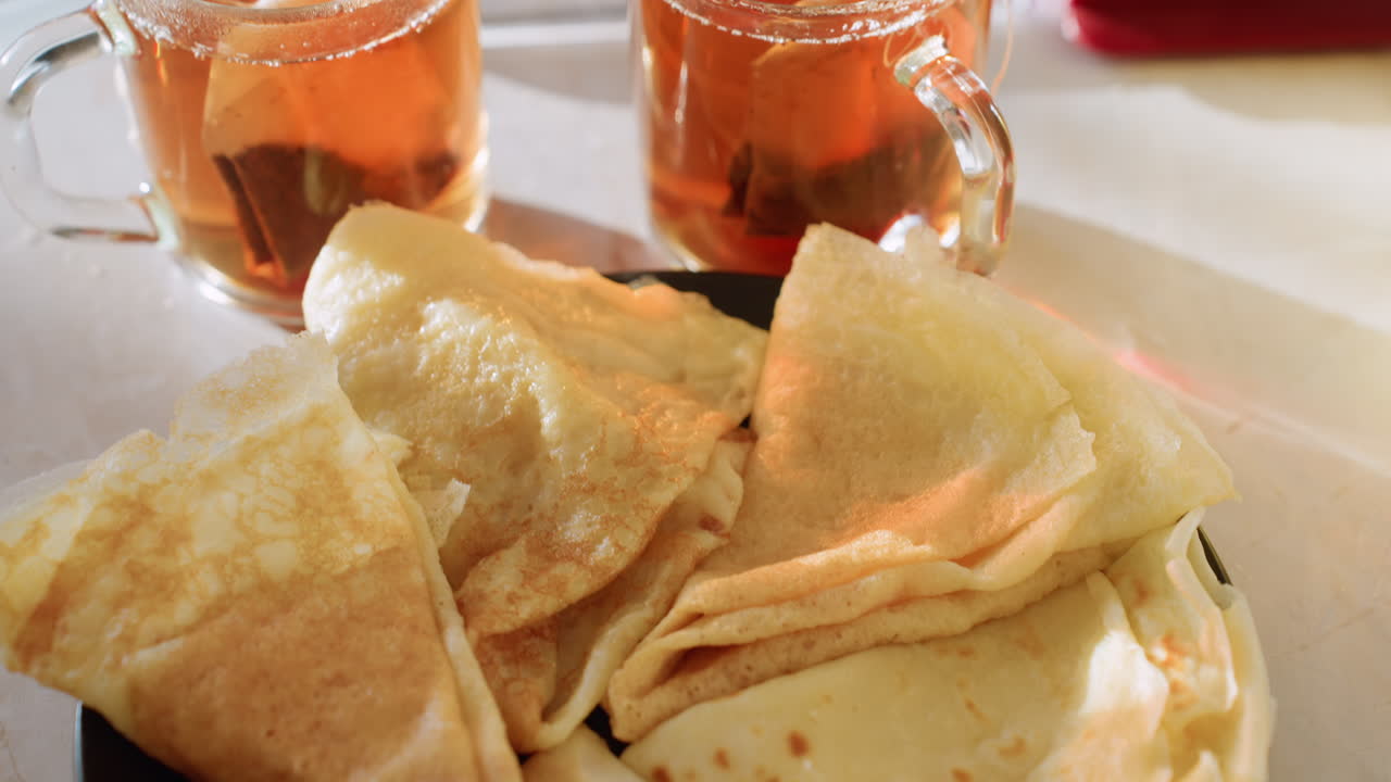 Close up of two steaming glass cups filled with amber tea as teabags steep, placed beside freshly made pancakes on kitchen counter in warm morning light
