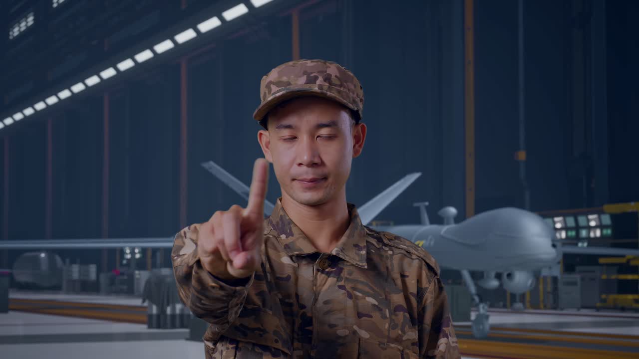 Close-up de Soldado Asiático Apontando Para Você e Desaprovando com o Sinal de 'Não' com o Dedo Indicador Enquanto Está de Pé com um Drone Militar Moderno em um Hangar de Manutenção