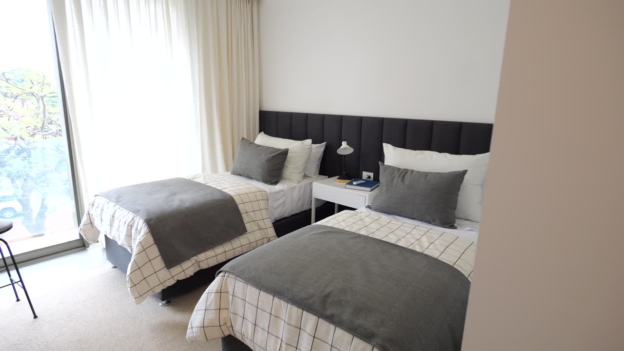 Two twin beds with gray sheets and pillows. The room is very clean and organized. The bed is next to a window, which lets in natural light