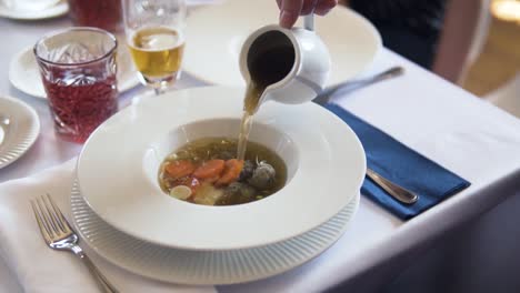 pouring a meatball soup into a plate on a set table with white tablecloth, slow motion