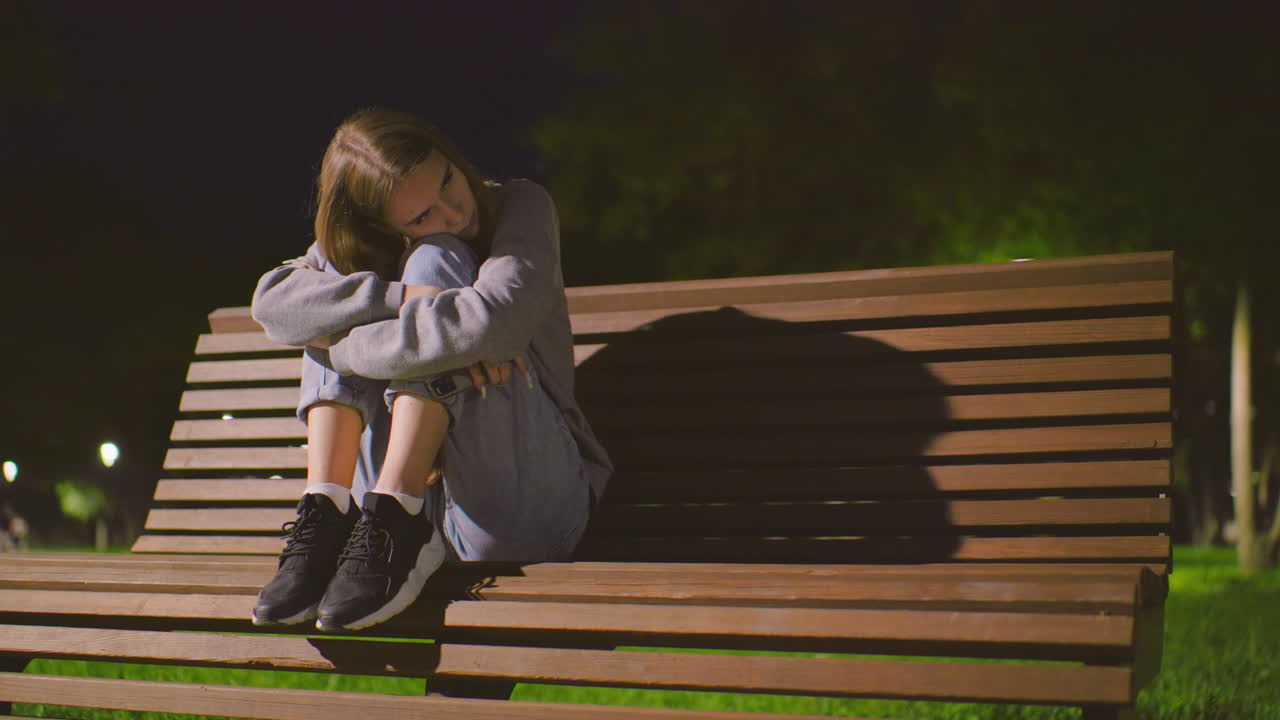 밤에 공원 벤치에 앉아있는 여인, 무을 둘러싼 팔, 다리에 머리를 고, 부드러운 조명 아래 멀리로 쳐다보고, 조용한 초록색으로 둘러싸여 벤치에서 그림자를 던집니다.