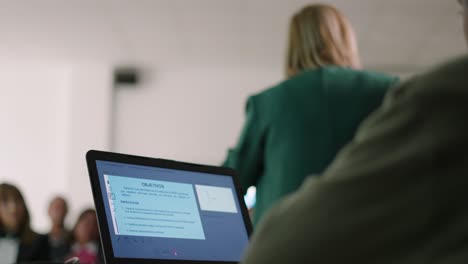 Hombre-Usando-Un-Cuaderno-Mientras-Una-Mujer-De-Atrás-Da-Clases-A-Un-Grupo-De-Personas