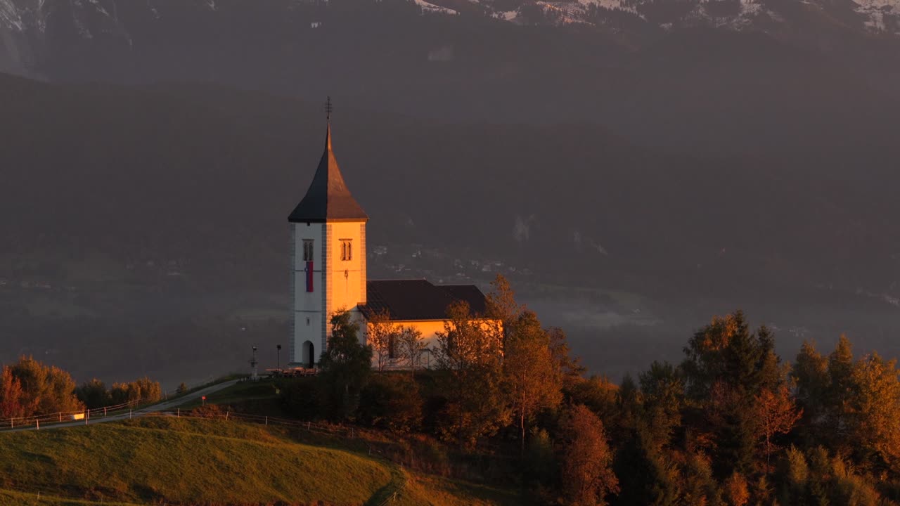 Aerial cinematic reveal of the Church of St. Primus and Felician on Jamnik Hill, Slovenia, with golden light over the Julian Alps highlighting the peaceful countryside and mountain scenery