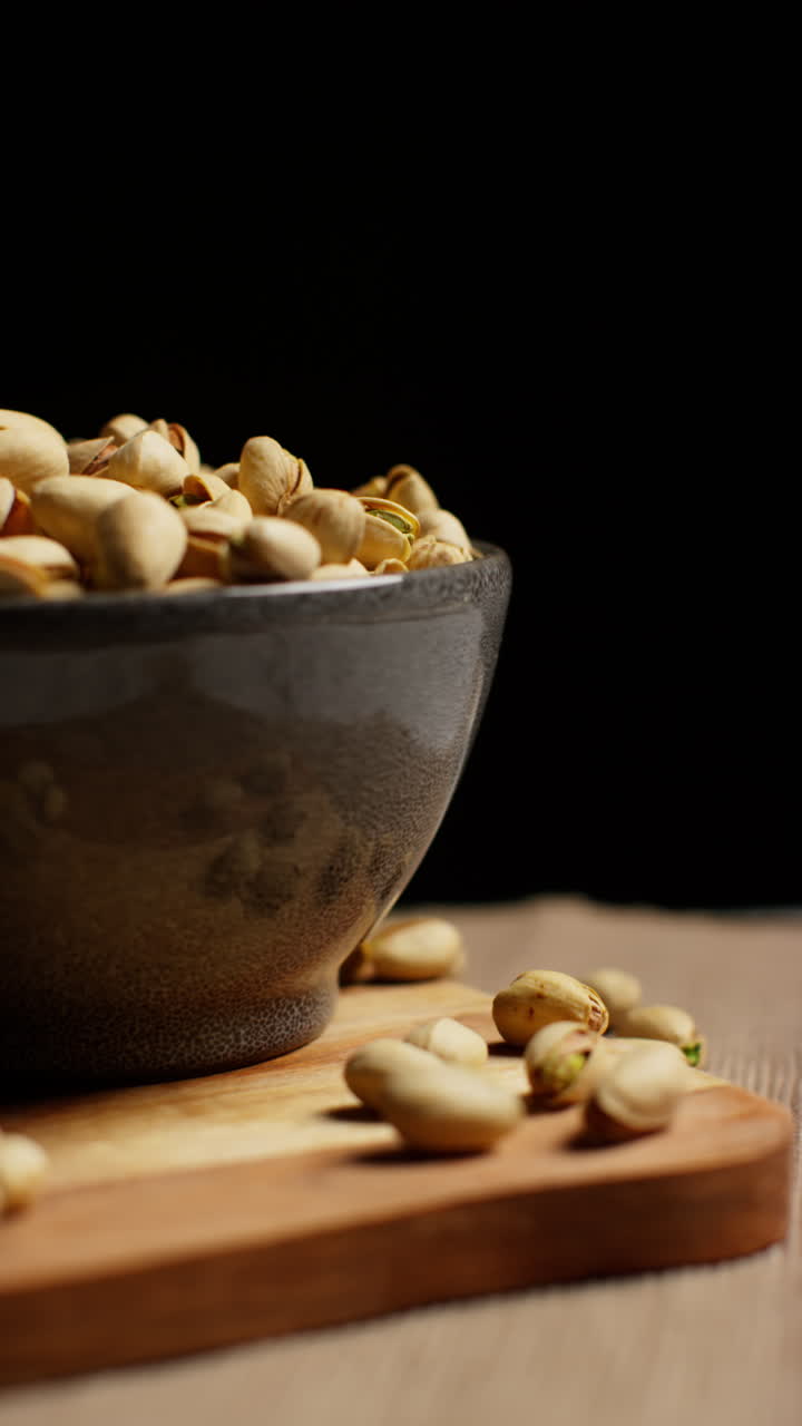 Vertical Video Close Up Studio Shot Of Bowl Of Pistachio Nuts In Shells Revolving Against Black Studio Background